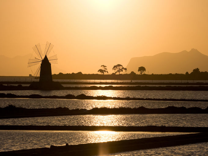 Silhouette di uno storico mulino a vento riflesso nelle vasche d'acqua allo Stagnone di Marsala durante un Tour Mozia con Autista Privato al tramonto.